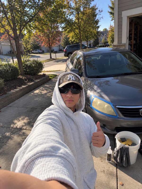 Shawn giving a thumbs up in a driveway next to a car with its yellowed headlights taped off and a bucket of sandpaper ready